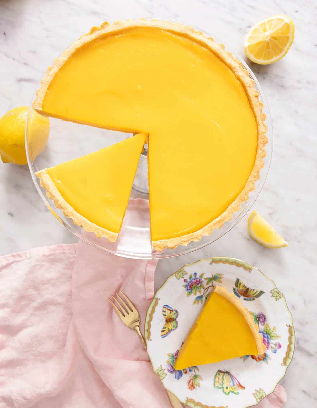 Overhead view of a lemon tart on a cake stand with a slice plated in front.