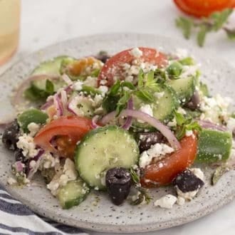 Greek salad on a spekled white plate next to a glass on white wine.