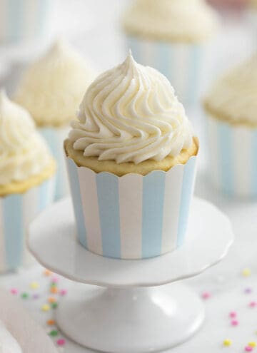 A photo of a vanilla cupcake on a cupcake stand with vanilla buttercream frosting beautifully piped on top.