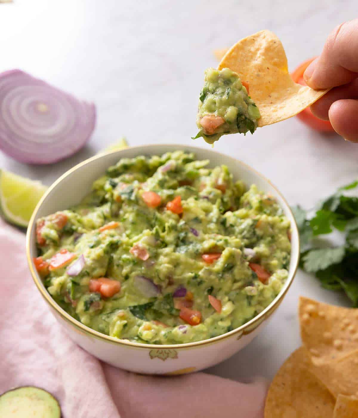 An overhead shot of a bowl of guacamole with tortilla chips inside of the bowl and ingredients scattered around the side.