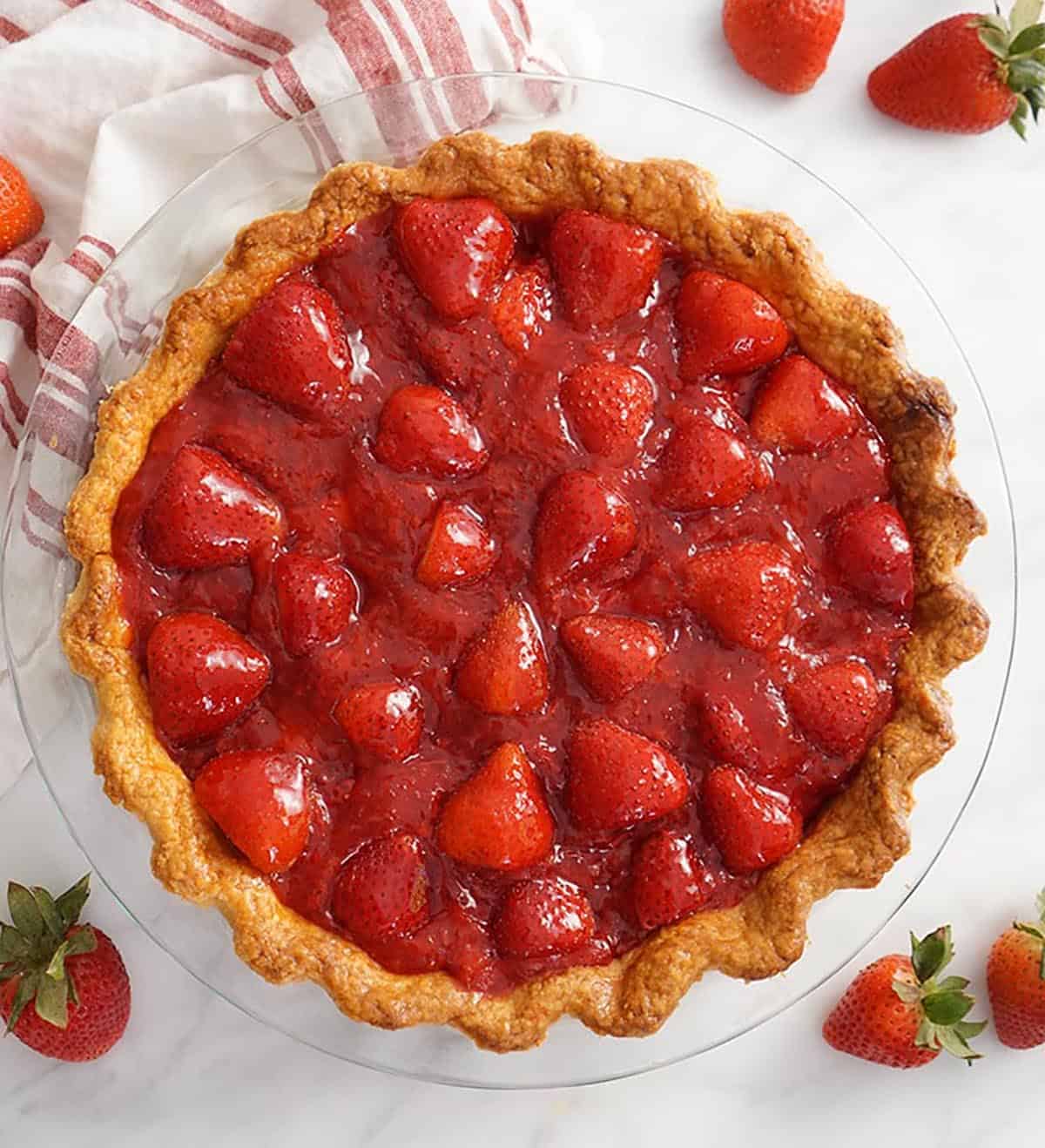 A top down shot of a whole strawberry pie on a marble table. Fresh strawberries scattered around.