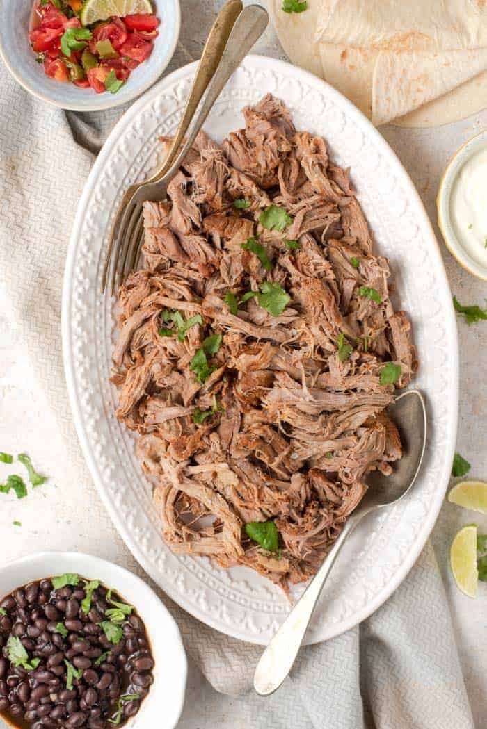 An overhead shot of instant pot pulled pork on a white platter. A bowl of black beans and salsa on the side.