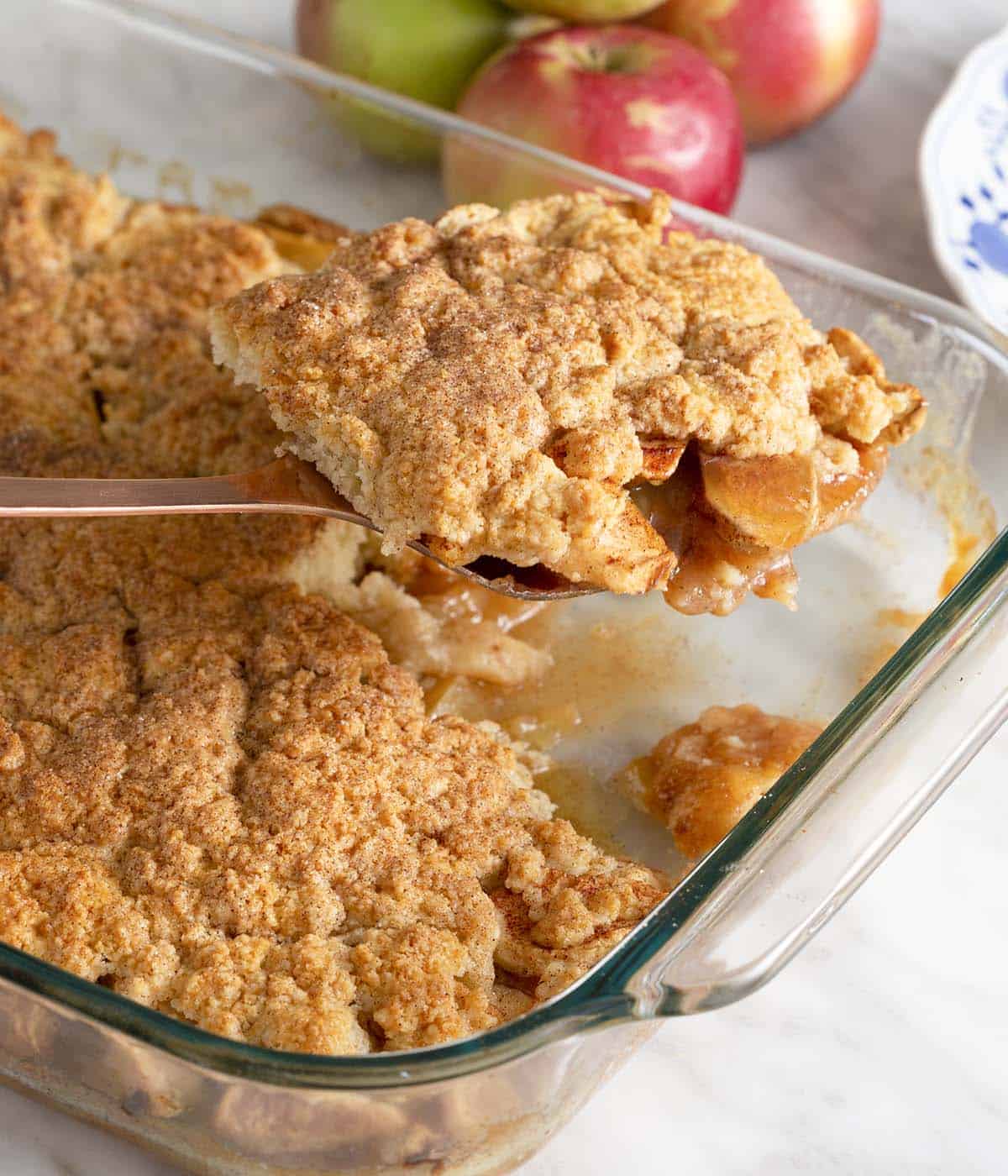 A serving spoon lifting up a serving of apple cobbler from a baking dish.