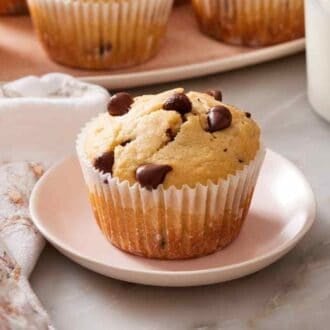 A chocolate chip muffin on a small plate next to a glass of milk and more muffins.