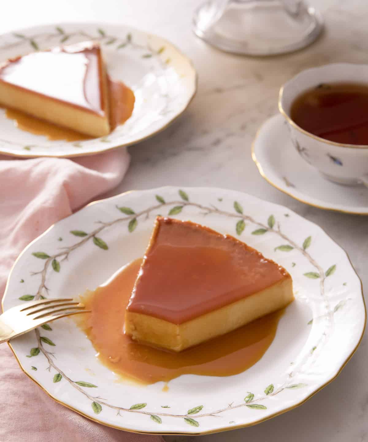 A piece of flan on a plate with a bite taken out with a fork. A mug of tea and another slice in the back.