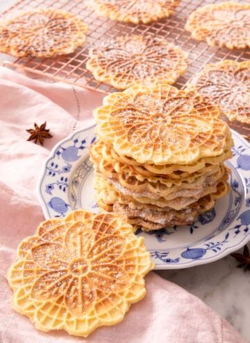 A plate with a stack of pizzelle cookies with more in the background on a cooling rack and one leaning on the plate.