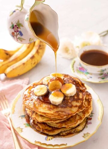 A jug pouring syrup over a stack of banana pancakes.