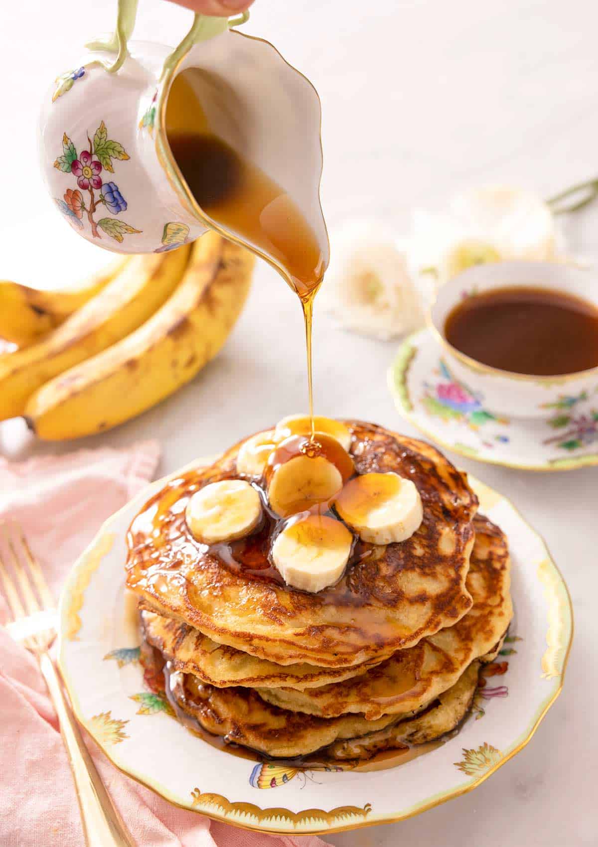 A jug pouring syrup over a stack of banana pancakes.