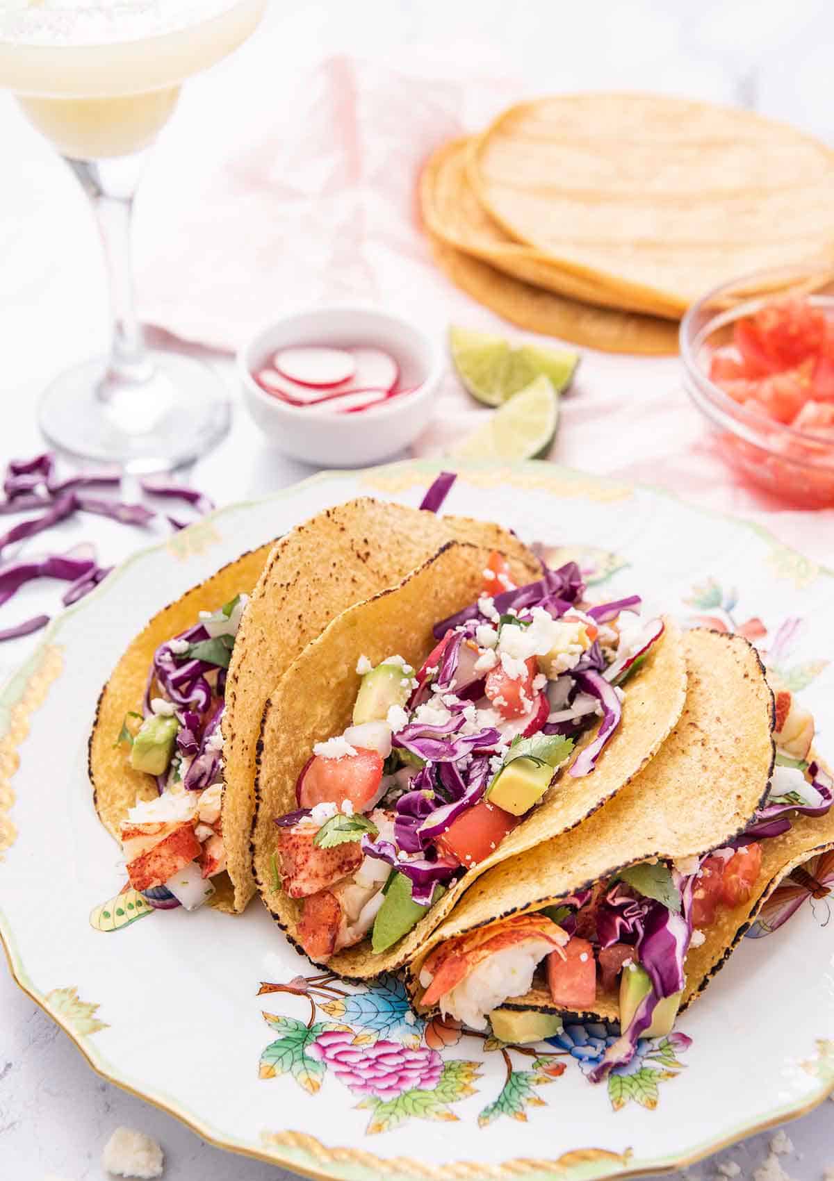 A plate of three lobster tacos in front of filling ingredients and a cocktail.