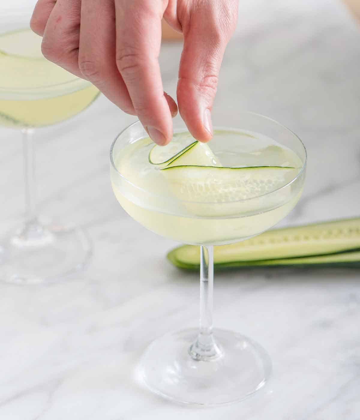 A hand placing in a sliced cucumber into a glass of cucumber martini.