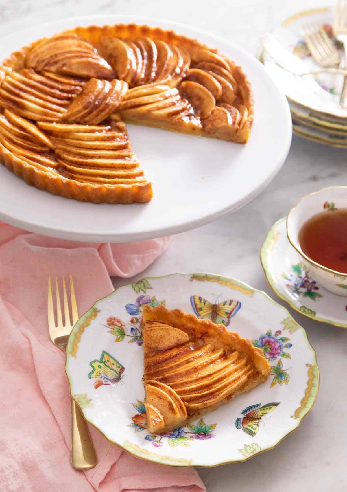 A serving of apple tart in a plate in front of the entire tart that it was cut out of.