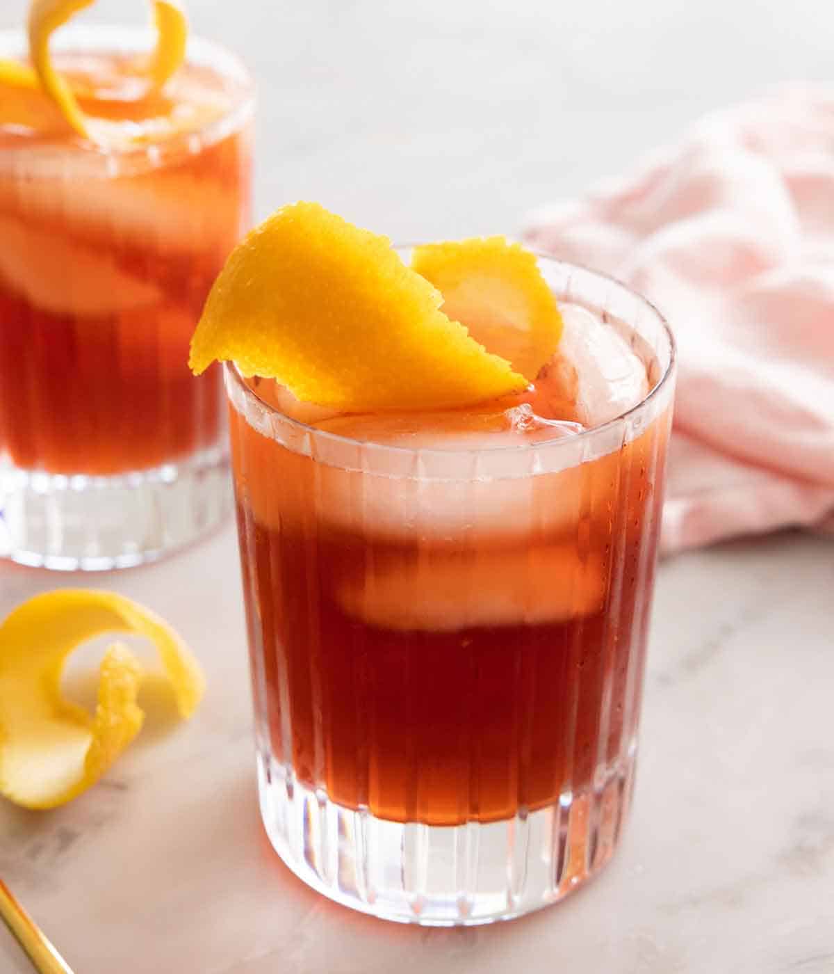 A glass of Negroni in front of a second glass and a pink linen napkin. Both has an orange peel garnish and one extra peel on the counter.