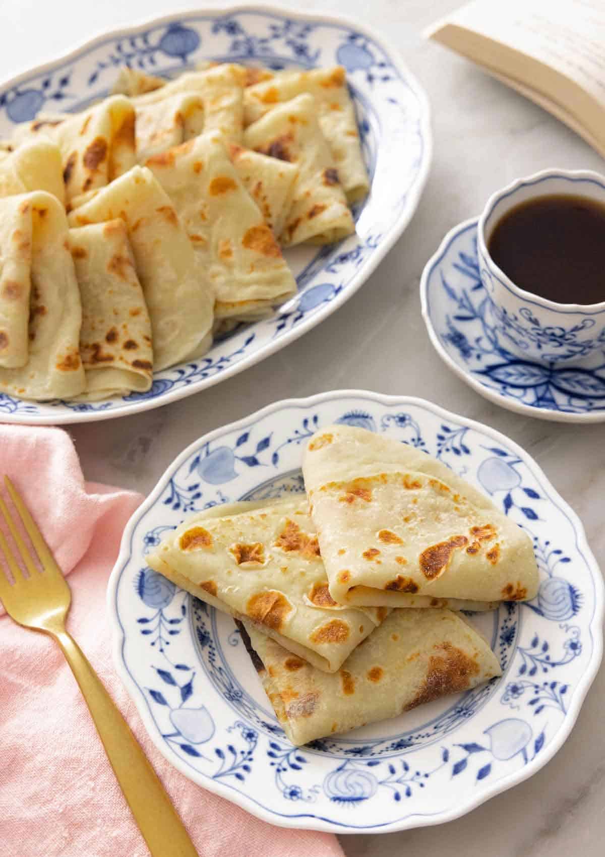 A plate with three folded lefse by a mug of coffee and a platter.