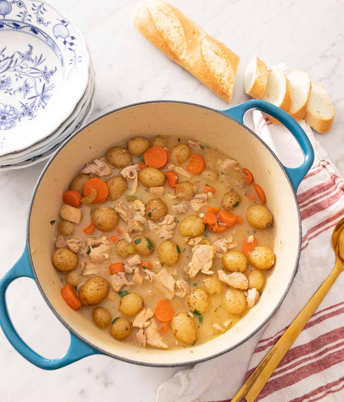 Overhead view of a Dutch oven containing chicken stew with sliced bread and a stack of plates nearby.