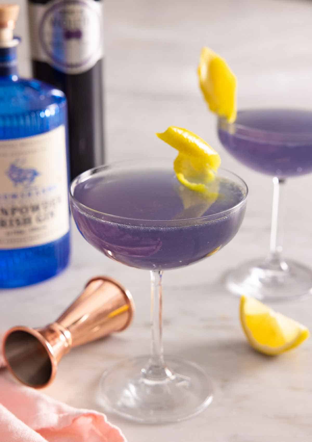 Two glasses of Aviation cocktail with lemon twist garnish by the bottles of liqueur in the back.