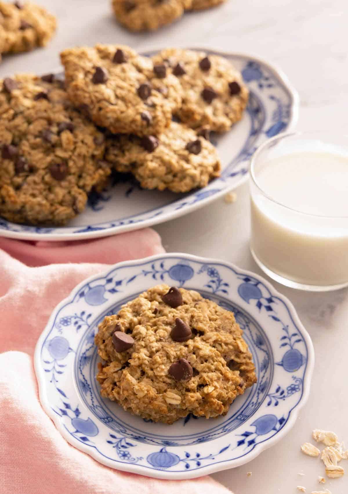 A plate with a banana oatmeal cookie by a glass of milk and a platter with more.