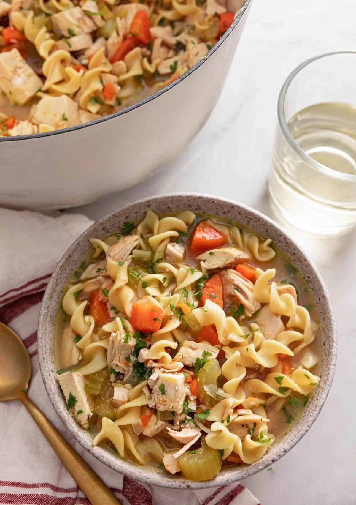 A bowl of chicken noodle soup by a glass of wine.