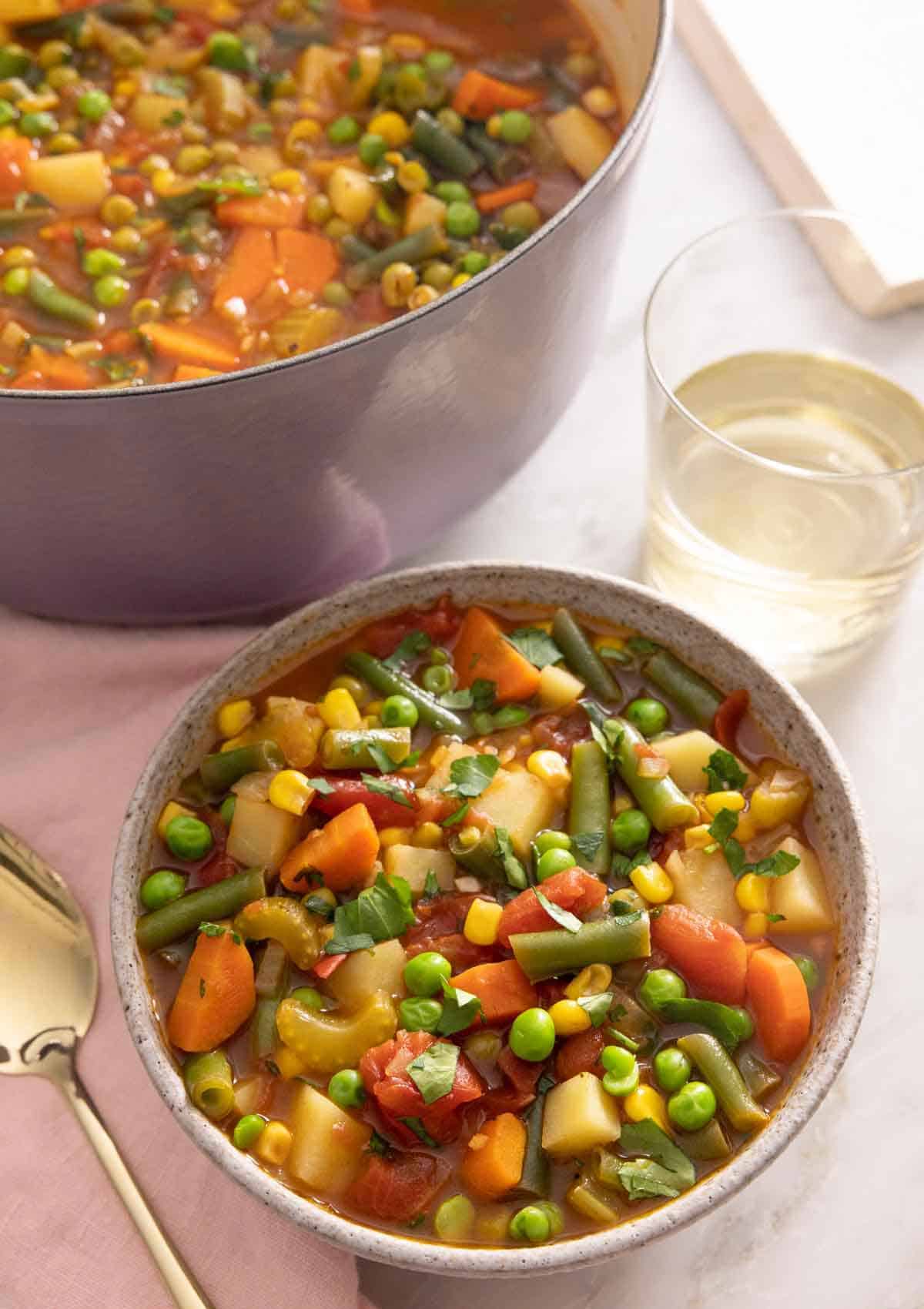 A bowl of vegetable soup by a glass of wine and pot of soup.