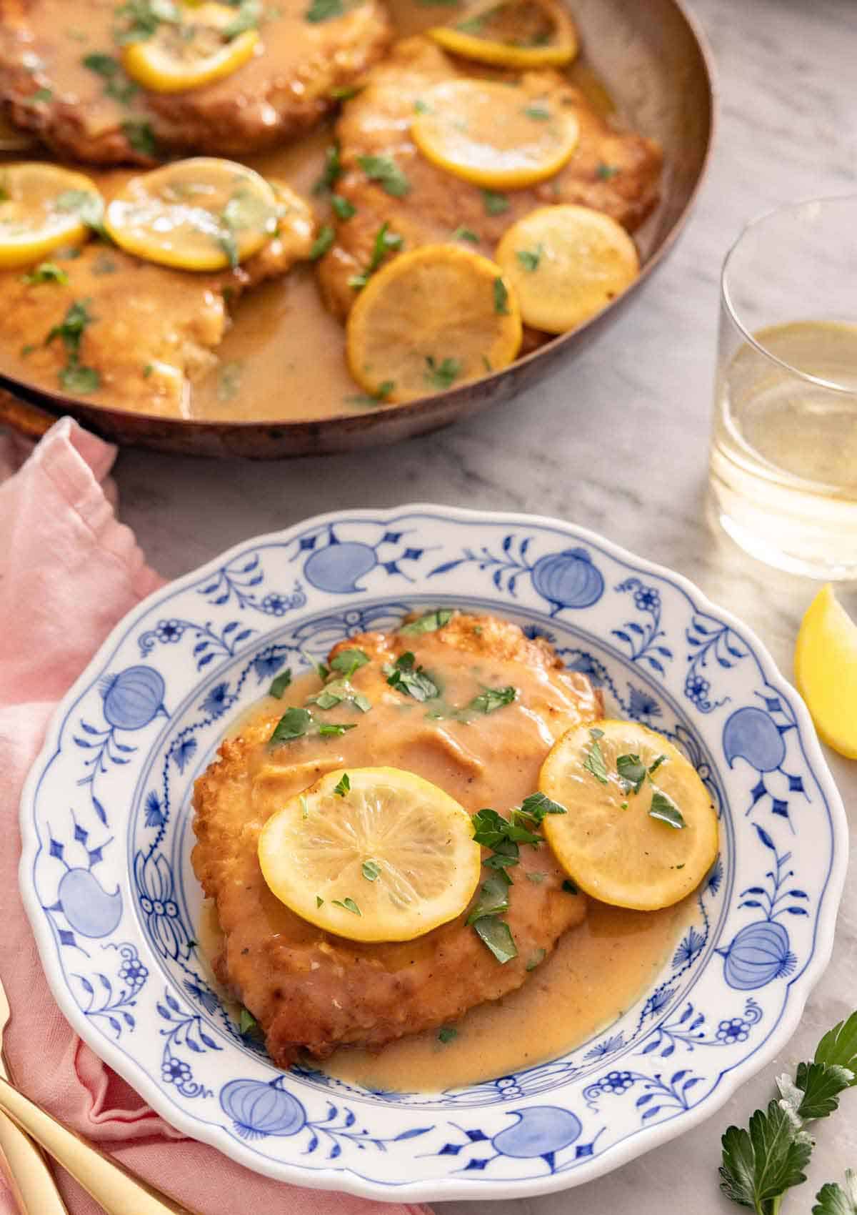 A plate of chicken Francese with a glass of wine in the background alongside a skillet.
