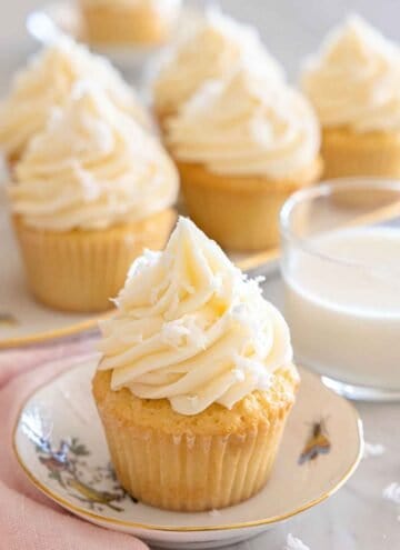 A plate with a coconut cupcake with more in the background along with milk.
