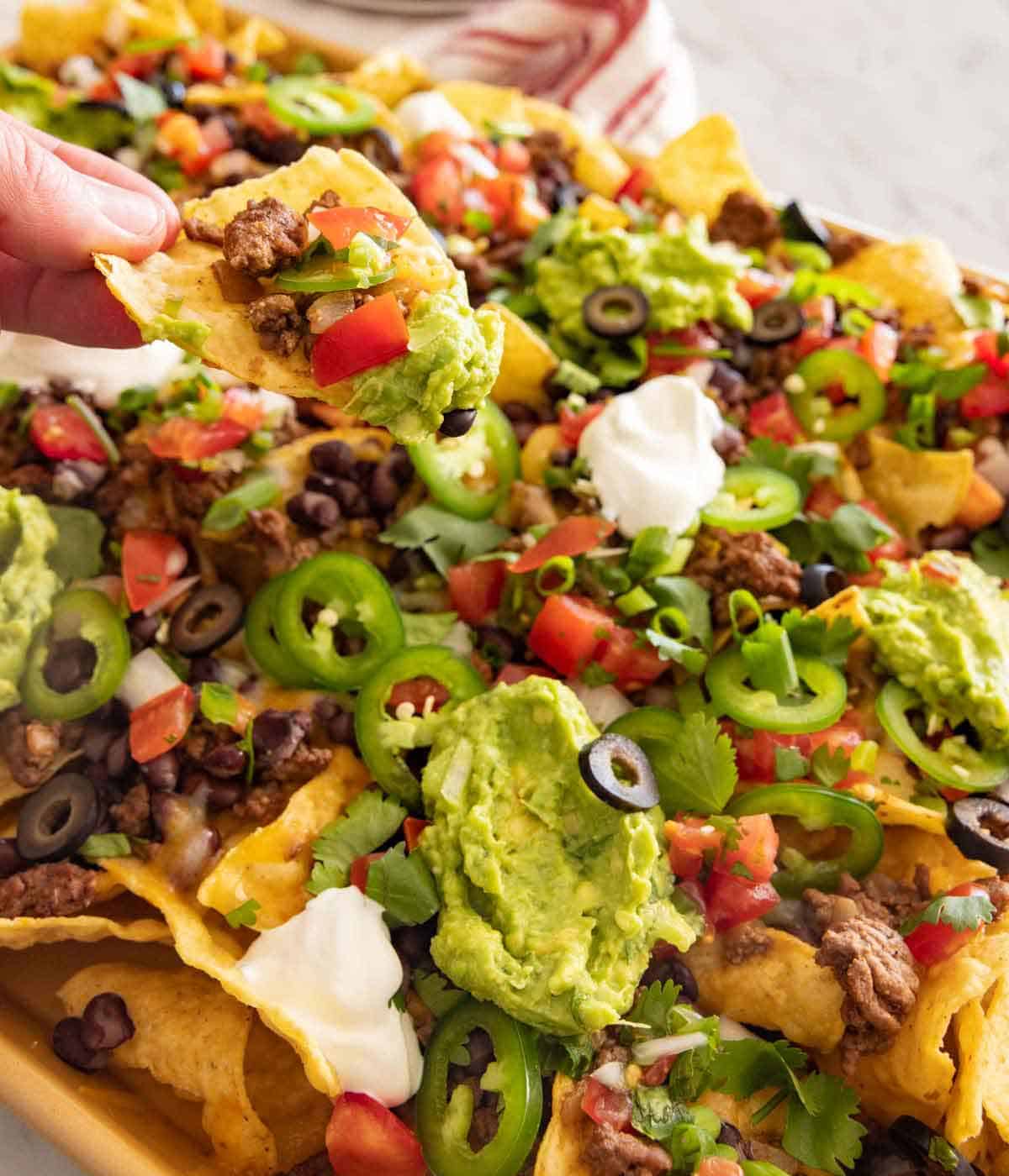 A tortilla chip being lifted out from a sheet pan of nachos.