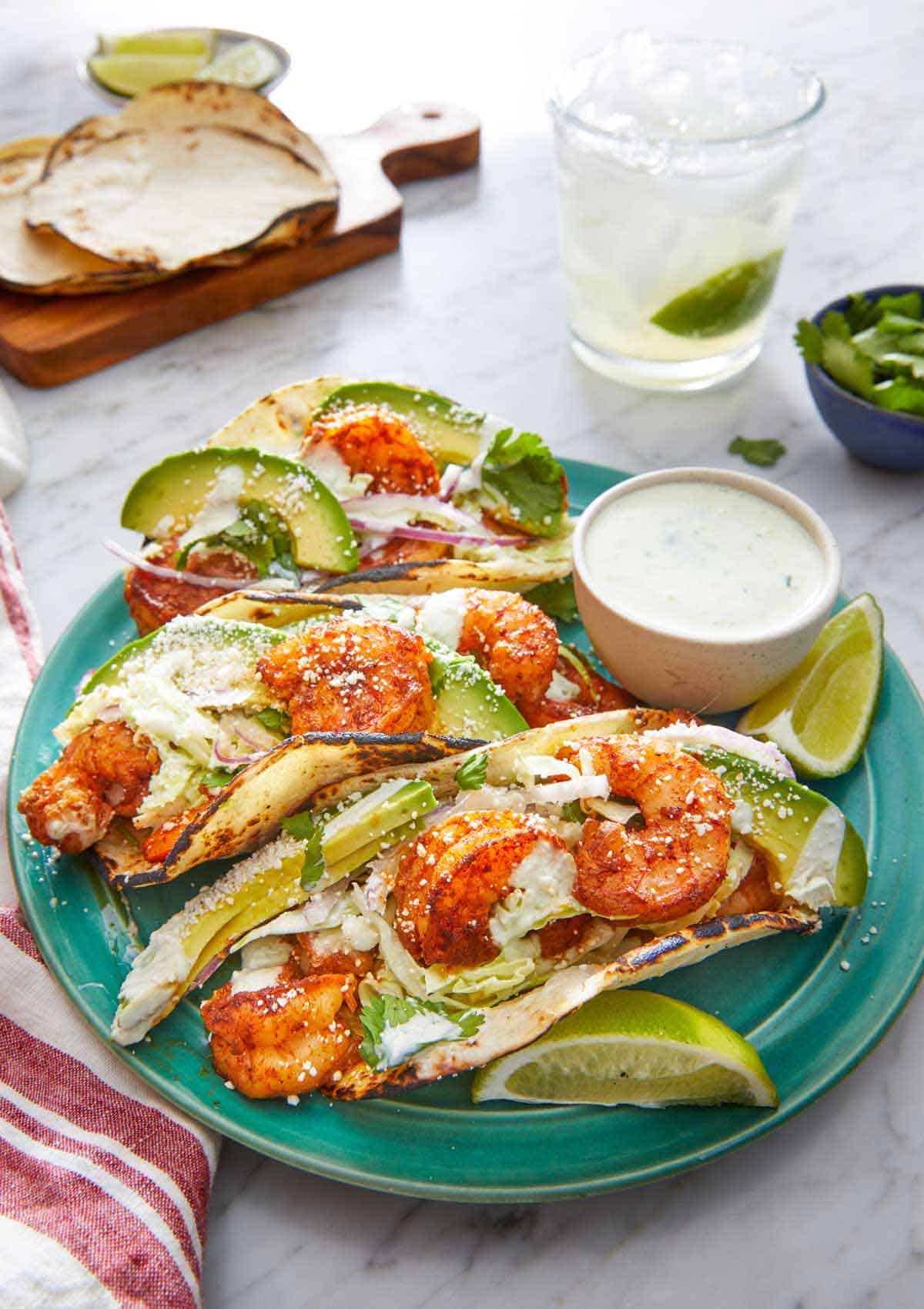 A plate with three shrimp tacos with a dip on the side with cotija cheese sprinkled on top of the tacos.