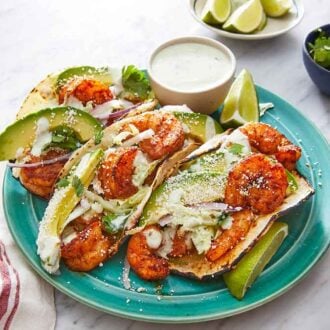 A plate with three shrimp tacos with a small bowl of dip with small bowls of garnishes on the side.