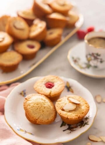 A plate with three financiers on a plate, one plain, one with a raspberry on top, and one with almonds on top.