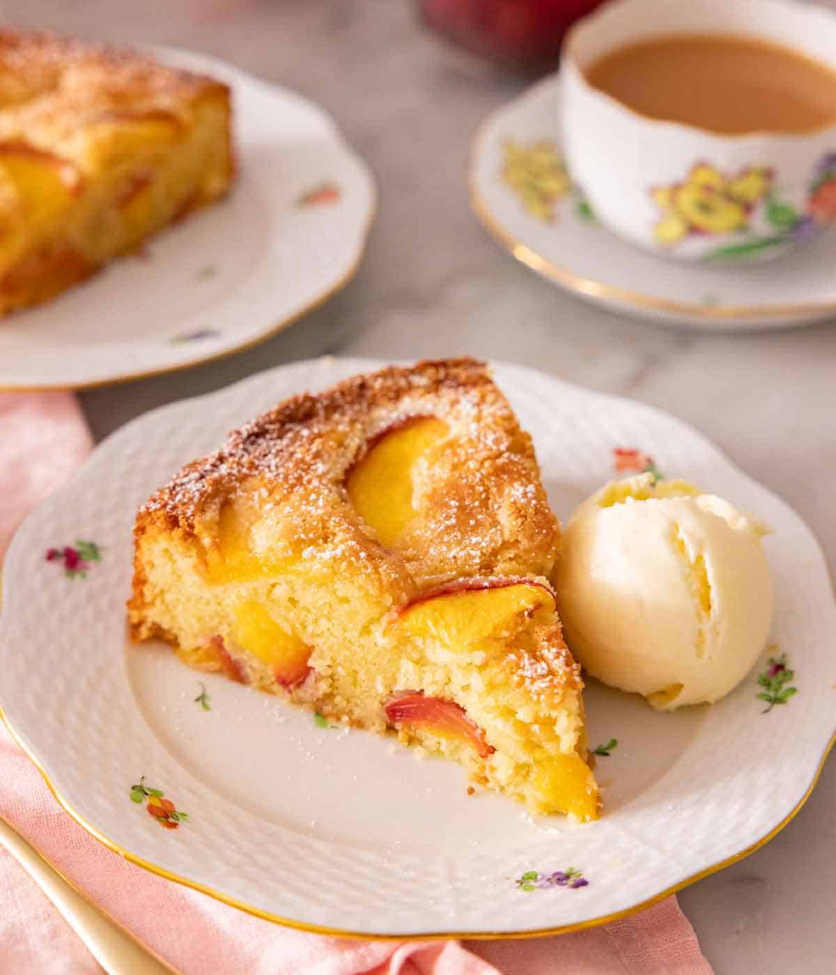 A plate with a slice of peach cake with a scoop over vanilla ice cream beside it with a cup of coffee in the background.