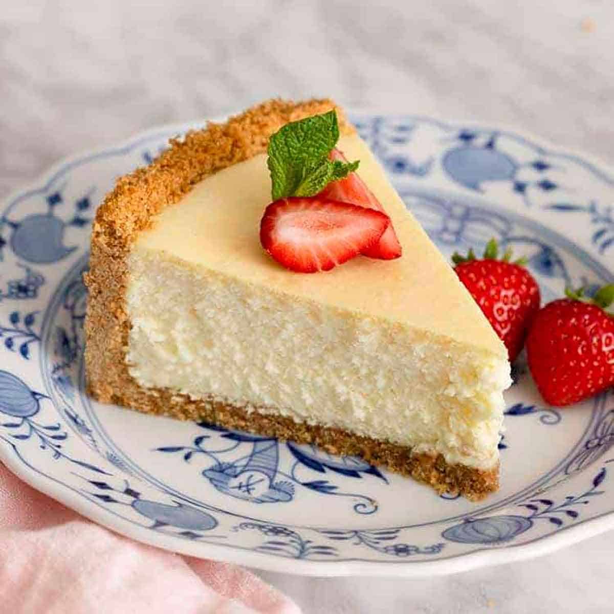 A slice of cheesecake with strawberry slices and fresh mint on top. Two strawberries on the side.