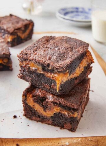 Two square pieces of caramel brownies stacked on top of each other on top of a lined cutting board.