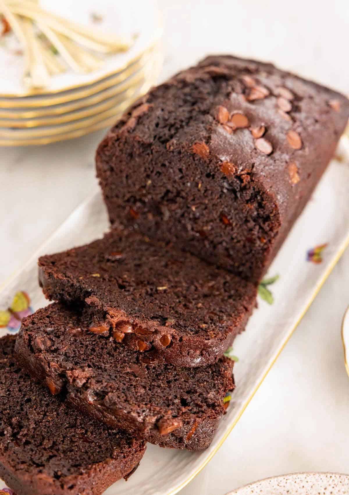 A platter with a loaf of chocolate zucchini bread with three slices cut in front.