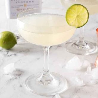 A glass of gimlet with ice, a jigger, and a whole lime behind it.