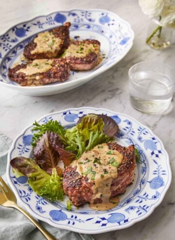 A plate of steak au poivre with a side of salad in front of a platter with three steak au poivre.