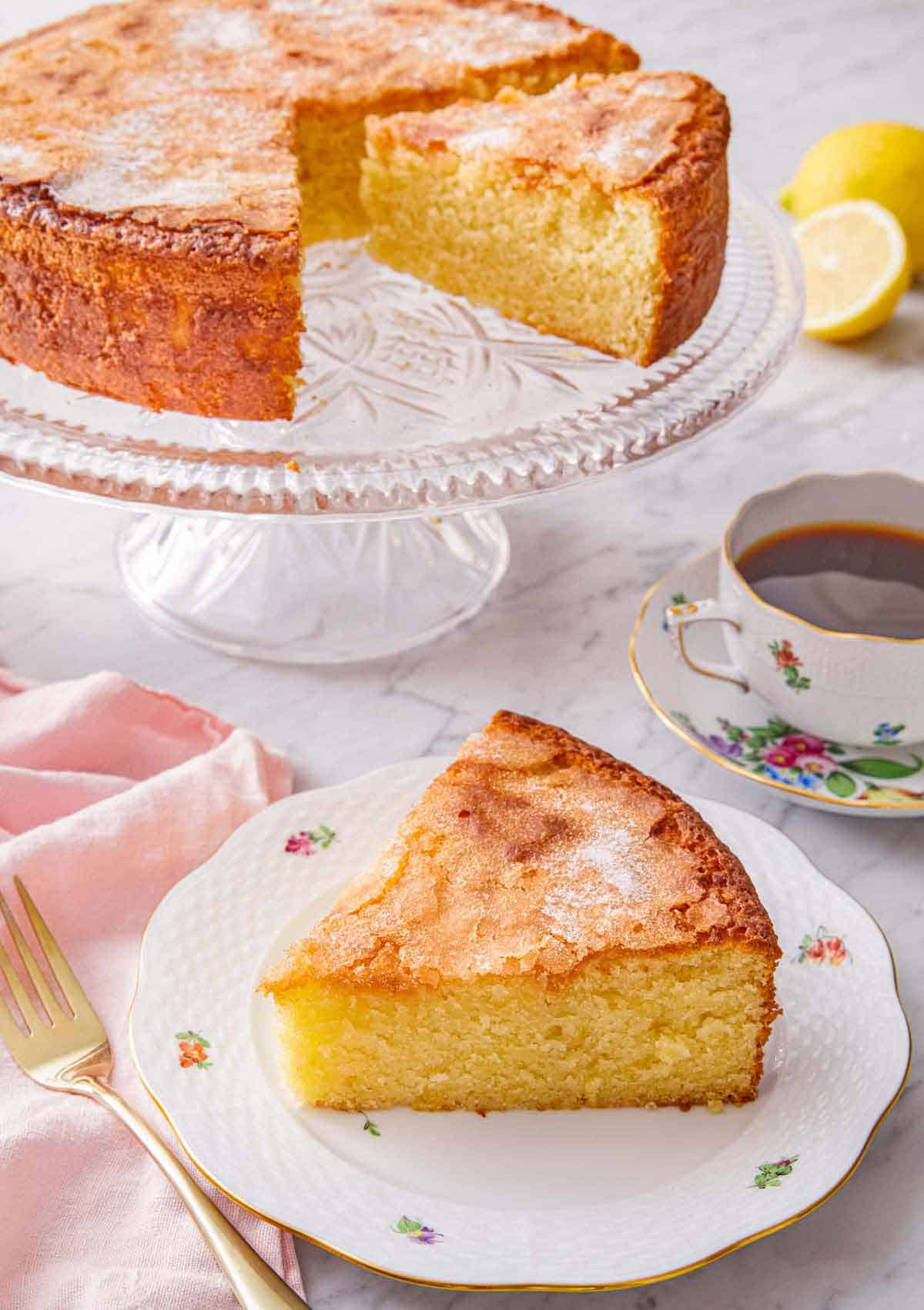 A slice of olive oil cake on a plate in front of the cut cake on a cake stand.