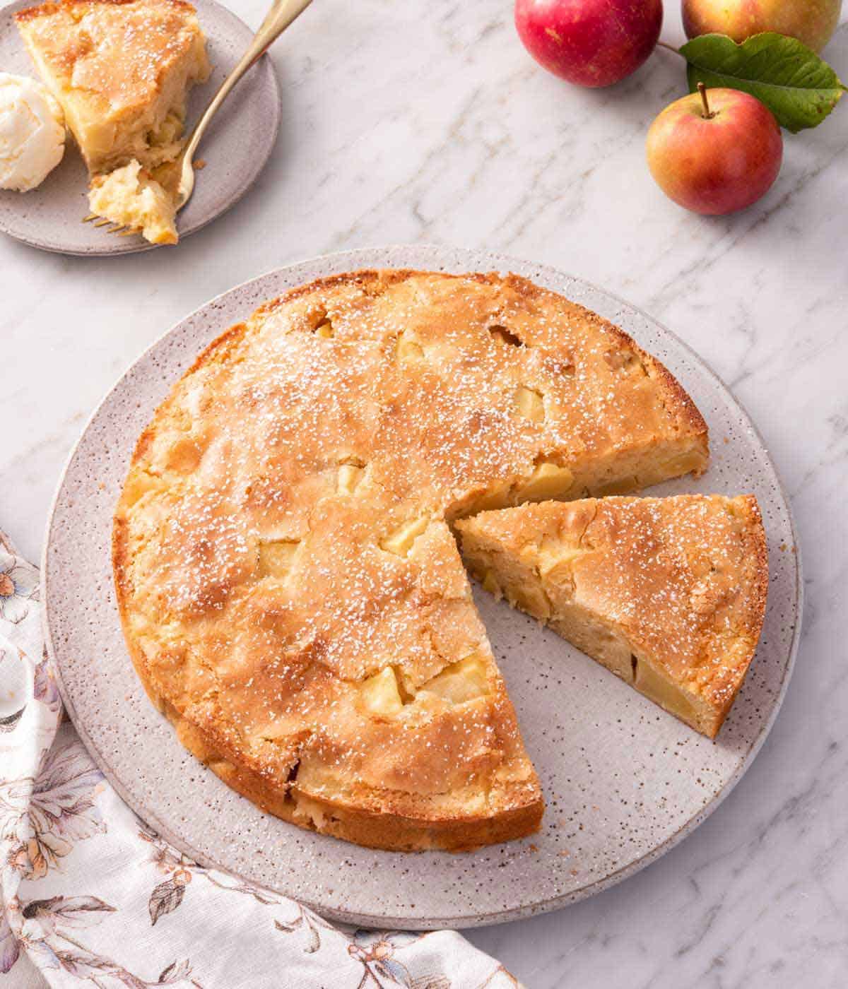 A slightly overhead view of a French apple cake with one slice removed and another slice cut and pulled forward.
