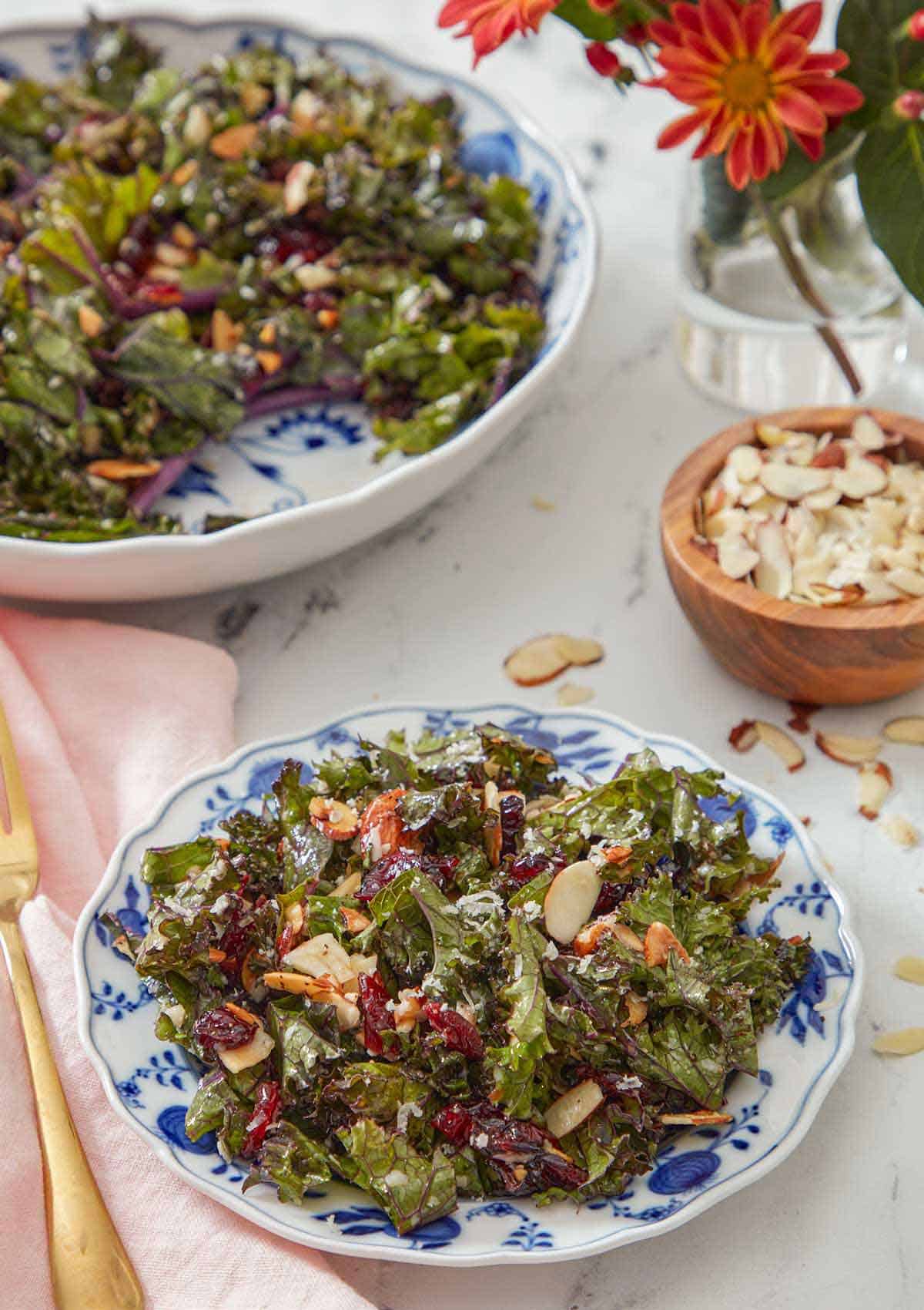 A plate of kale salad with a small bowl of almonds and serving bowl of salad in the back.