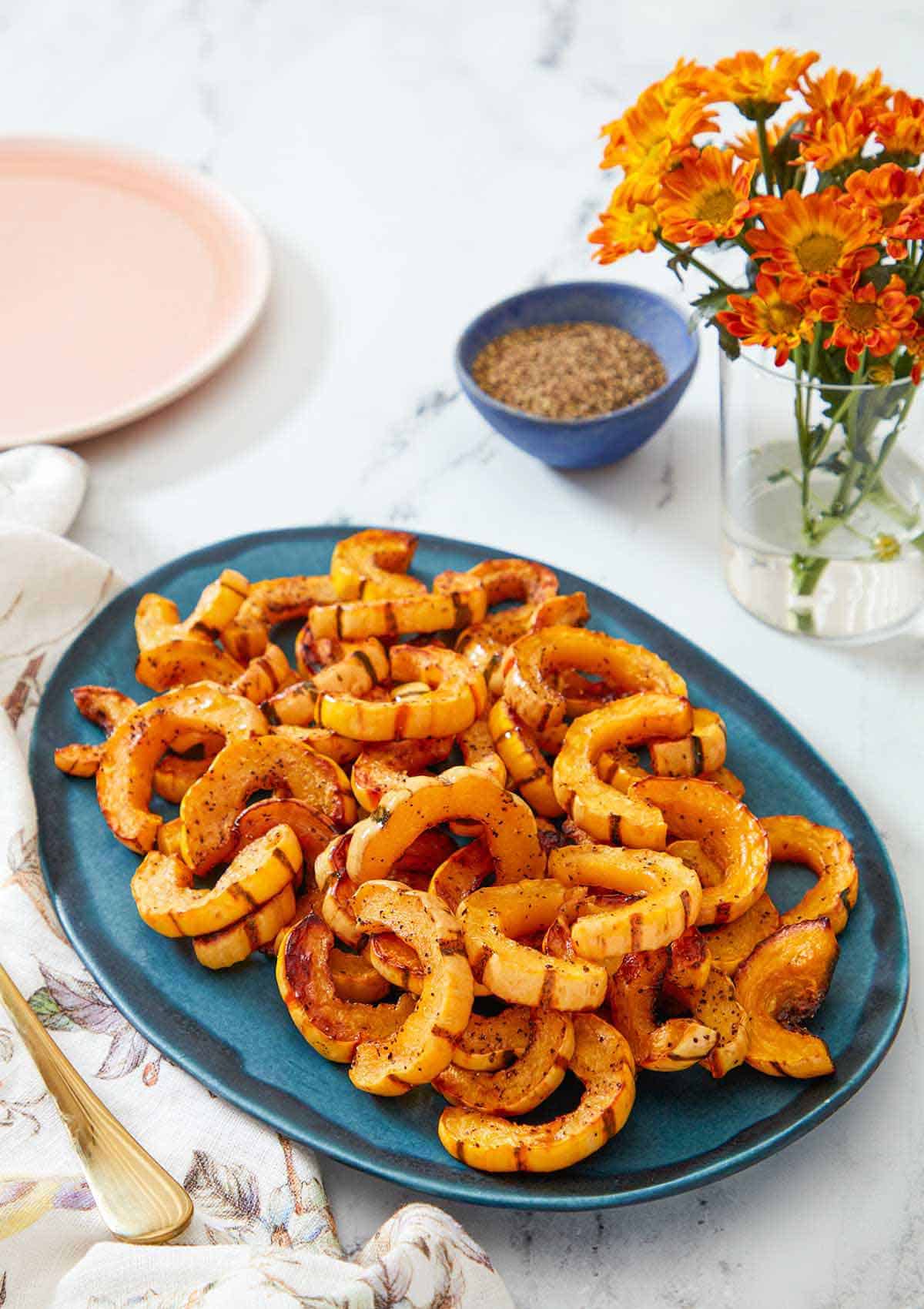 A blue serving platter with roasted delicata squash. A vase of flowers and a pinch bowl of pepper in the background.