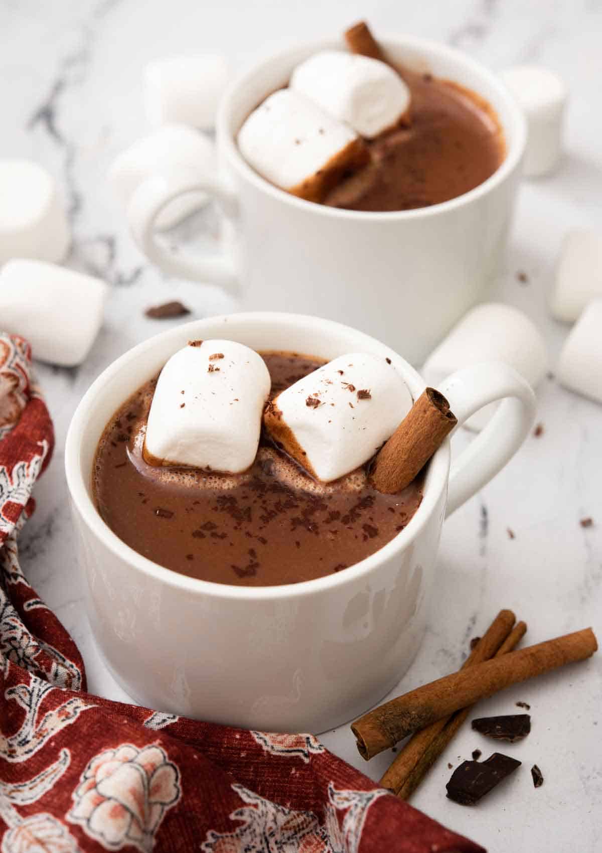 Two mugs of Mexican hot chocolate with marshmallows and cinnamon sticks inside. More garnish scattered around the counter.