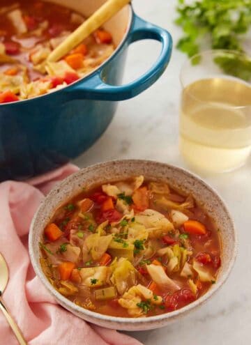 A bowl of cabbage soup with a pot in the back and glass of wine.