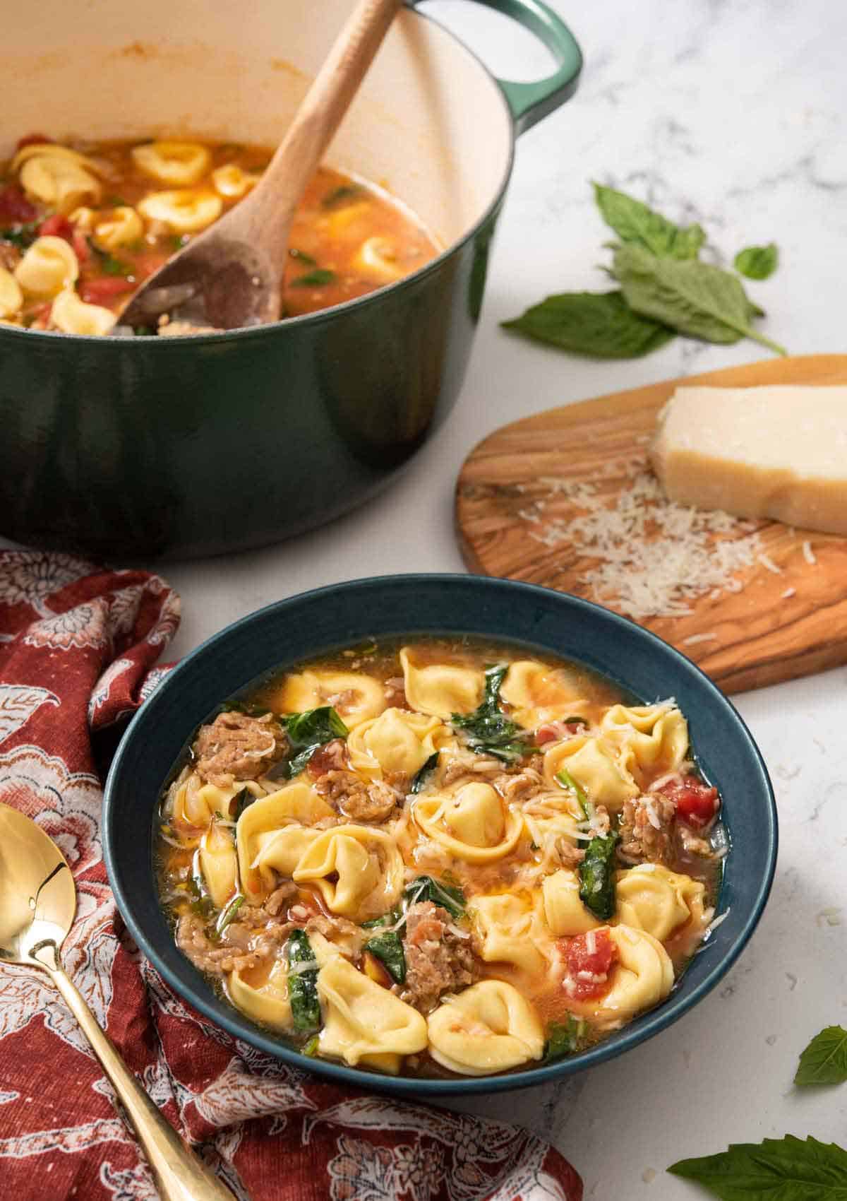 A bowl of tortellini soup with a pot in the back and grated parmesan on a cutting board.