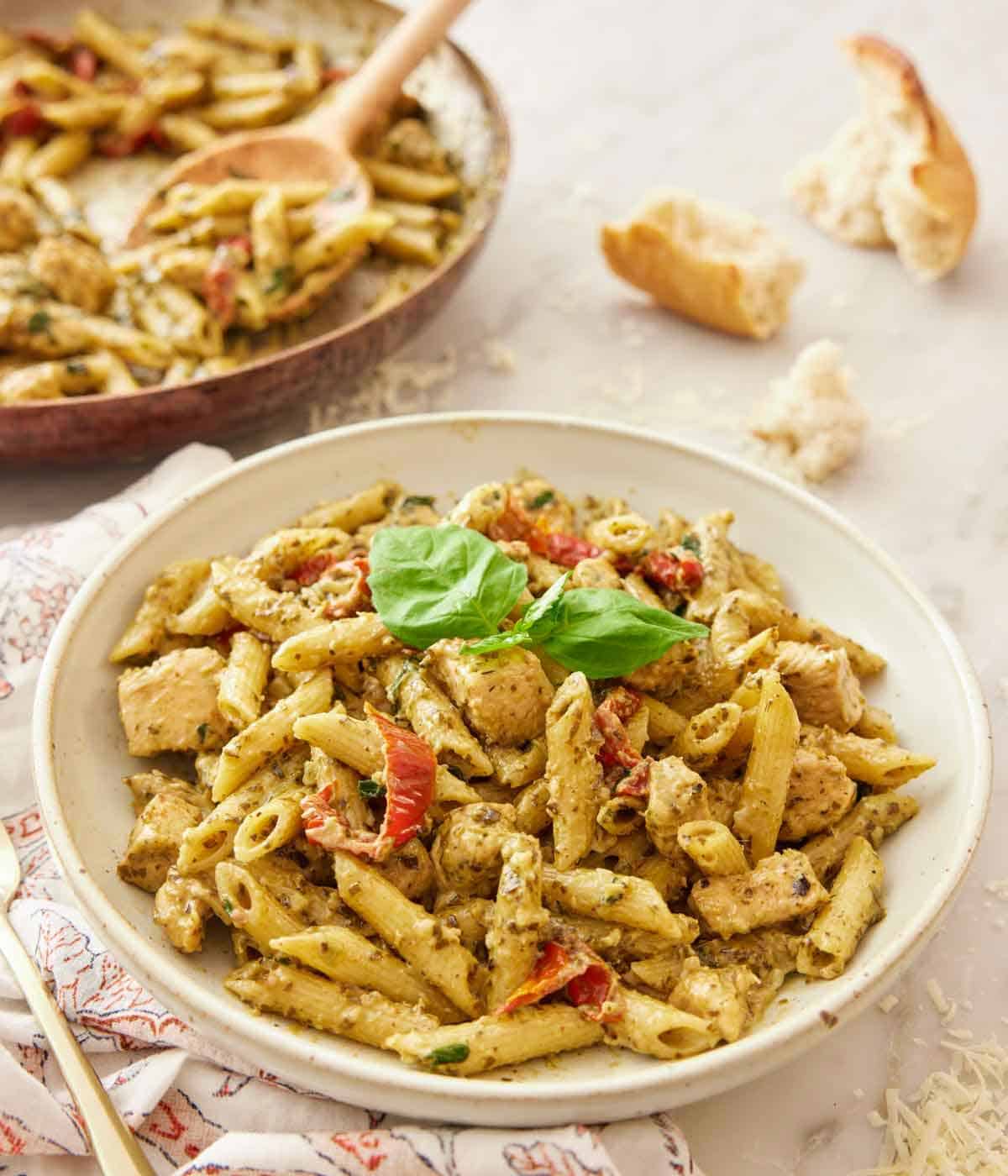 A plate of chicken pesto pasta with fresh basil on top with a skillet with a spoon scooping more pasta in the back.