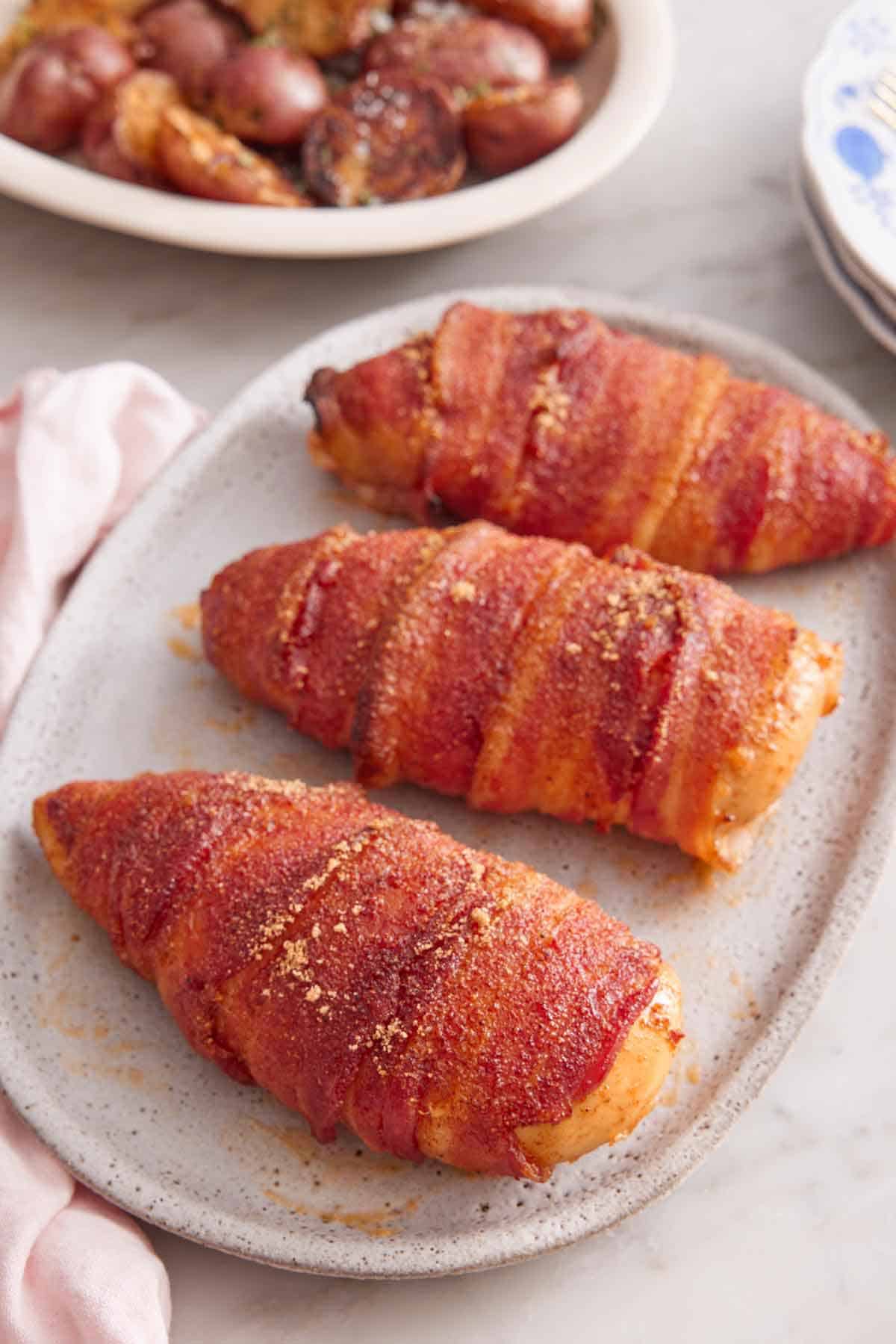A platter with three bacon wrapped chicken breasts with a plate of potatoes in the back.