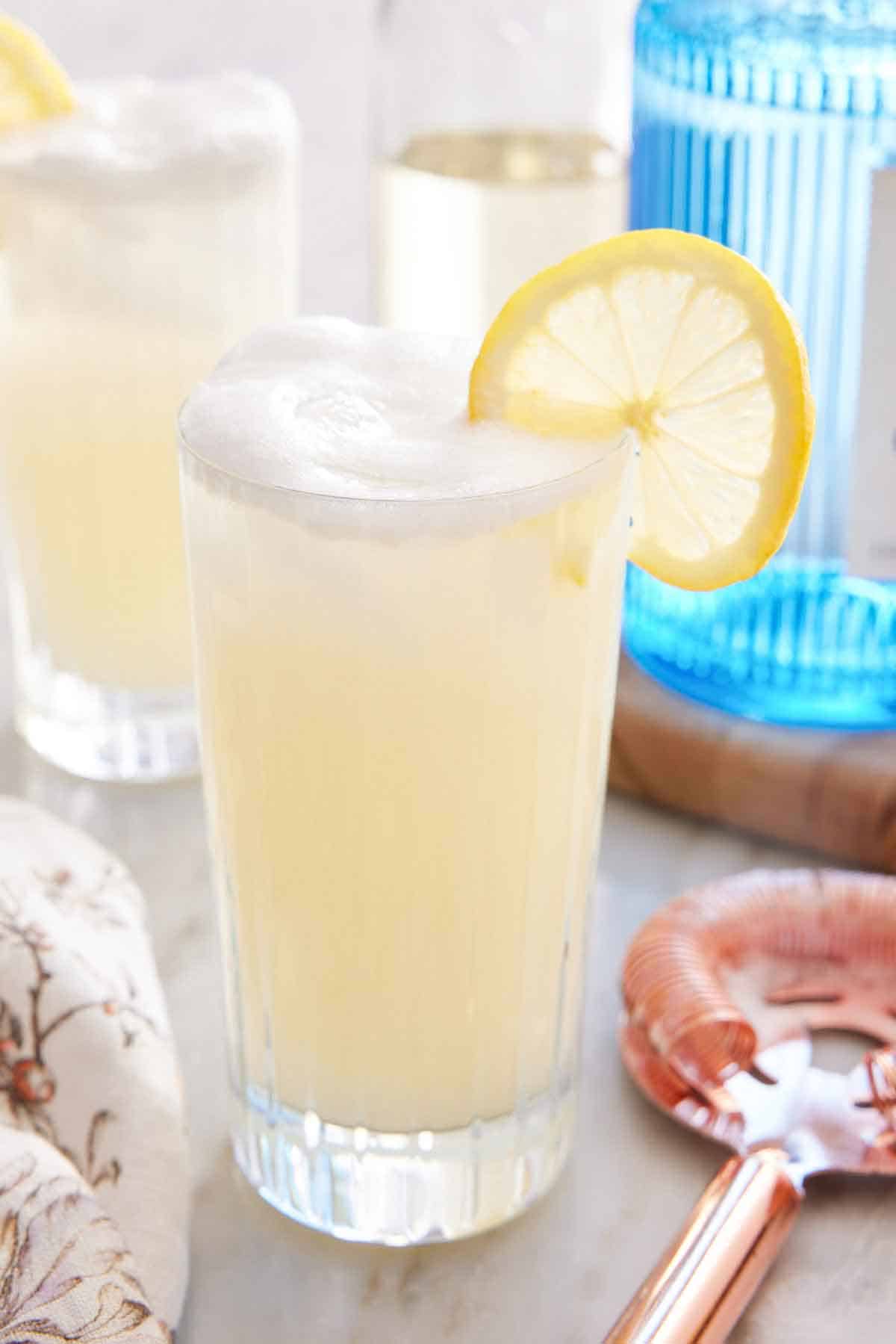 A glass of Gin Fizz with a slice lemon garnish on the rim with a cocktail strainer beside it and another glass of Gin Fizz and gin in the background.