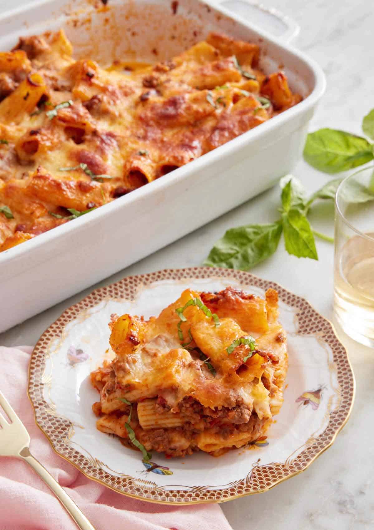 A plate with a serving of baked rigatoni with the baking dish in the back and some basil and wine to the side.