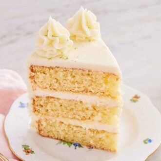 A slice of key lime cake with two dollops of frosting and lime zest on top. Glass of milk, fork, and cut limes beside it.