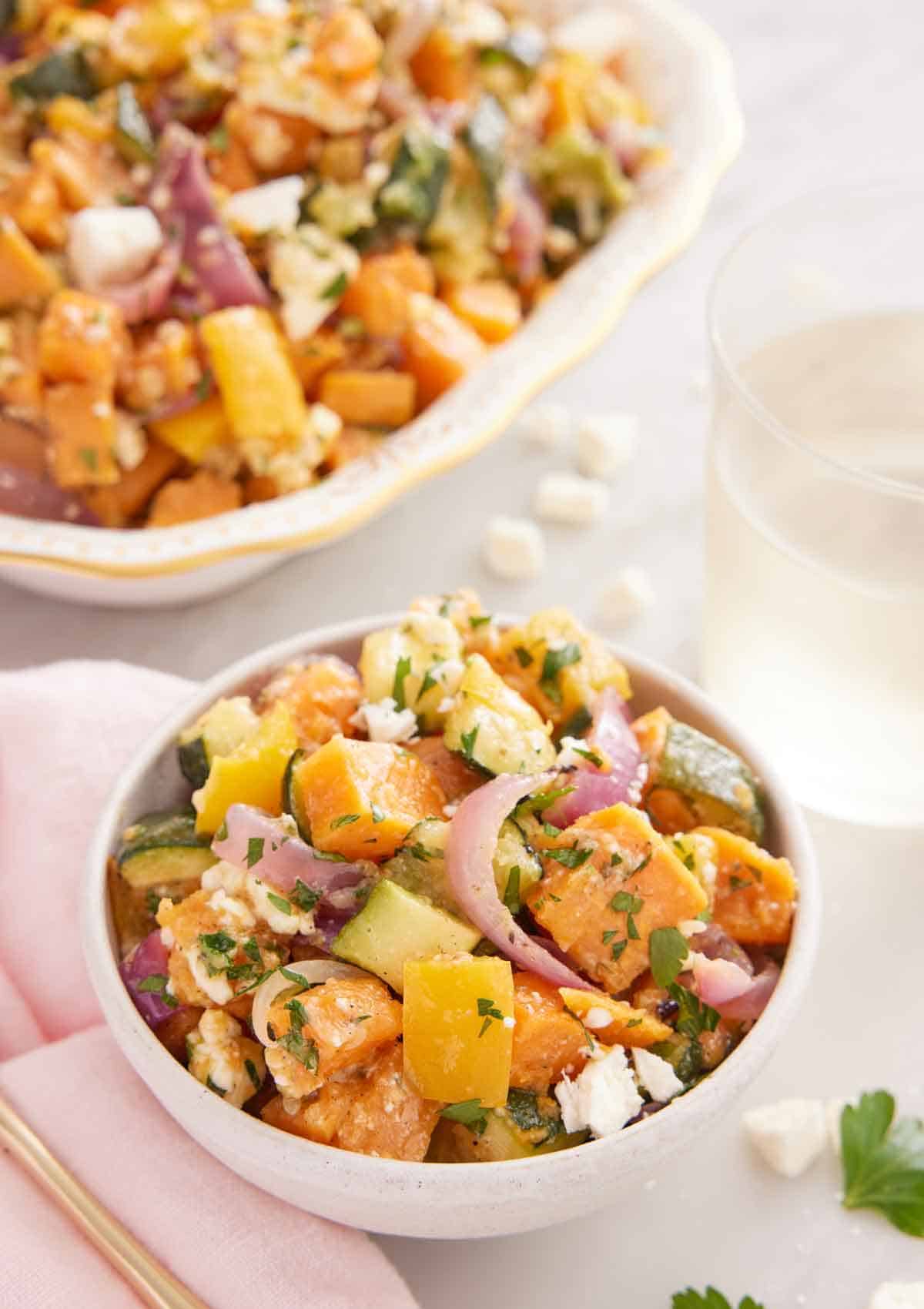 A bowl of roasted vegetable salad with a glass of wine and platter in the back.