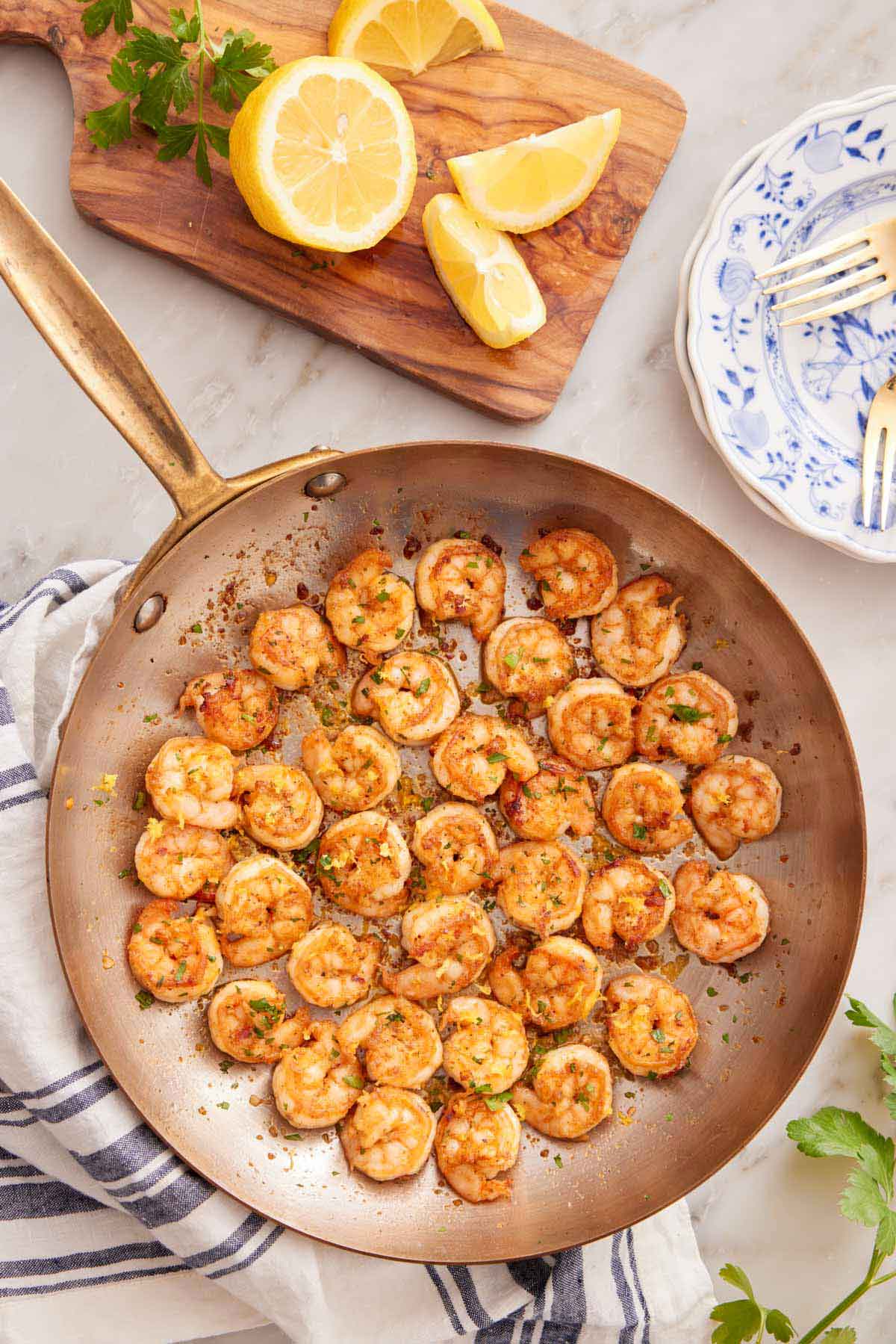 Overhead view of a skillet of sautéed shrimp with a cutting board with lemons and parsley beside it.