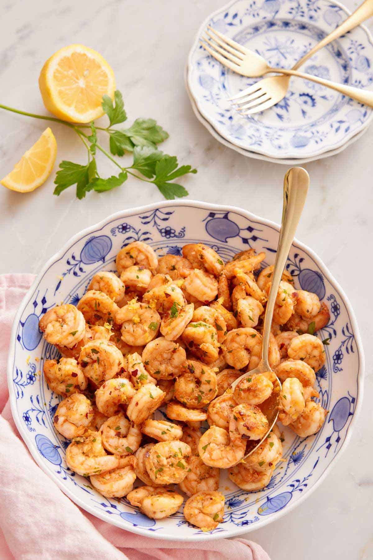 A large serving plate of sautéed shrimp with a spoon with some parsley, lemon, stack of plates, and forks in the back.