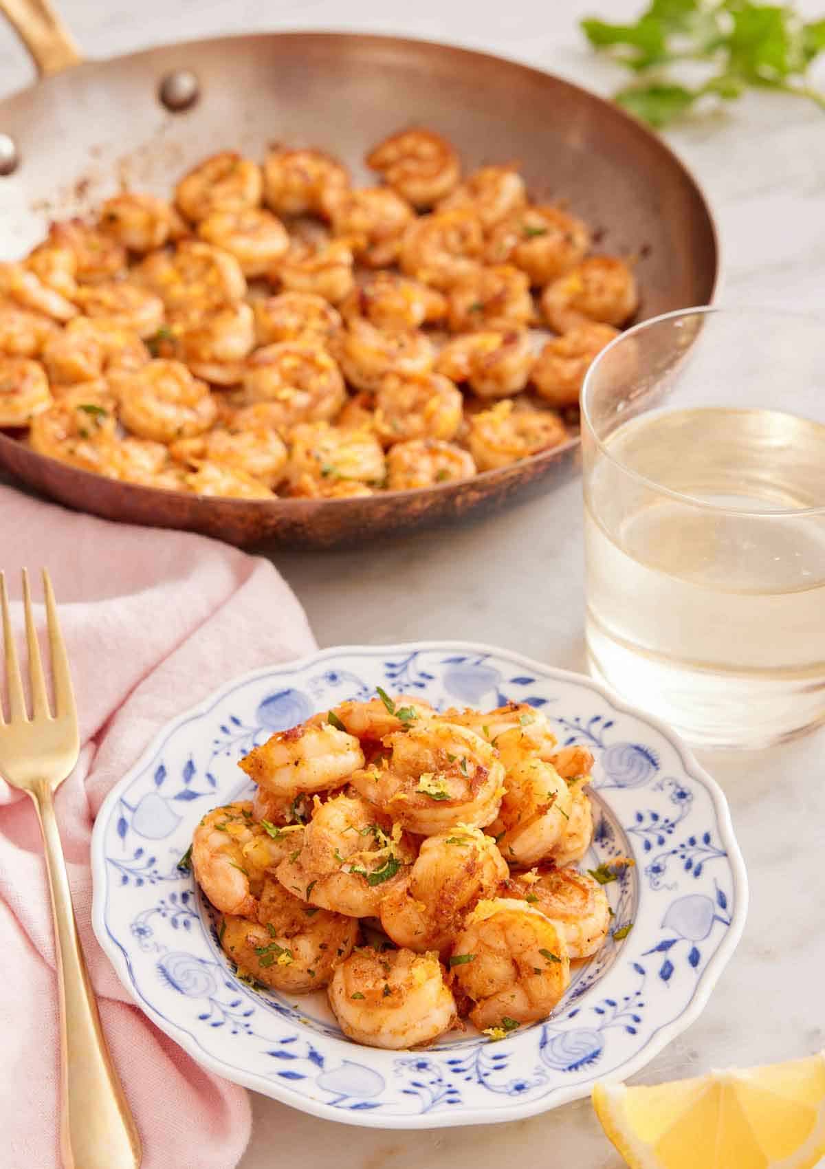 A plate of sautéed shrimp with a glass of wine and skillet of shrimp in the back.